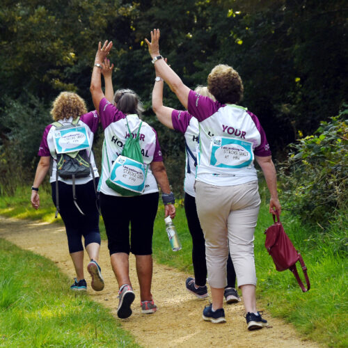 Four ladies taking part in a charity walk. They are walking along a path with their hands in the air, Frimley Health Charity t-shirts with 'walking in memory of' signs attached to them.