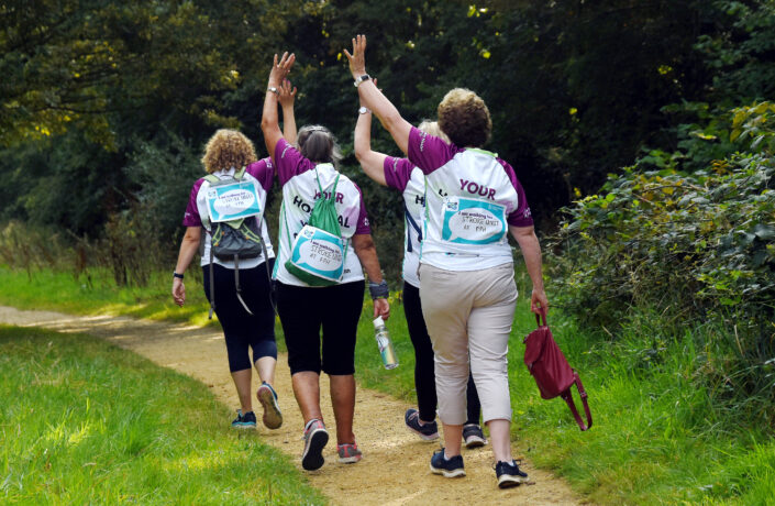 Four ladies taking part in a charity walk. They are walking along a path with their hands in the air, Frimley Health Charity t-shirts with 'walking in memory of' signs attached to them.